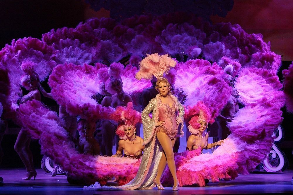 Woman in sparkly outfit and feathered headdress performs on stage with dancers, surrounded by large pink flower prop.