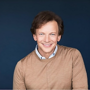 Smiling man with light brown hair in a tan sweater, standing in front of a dark blue background.