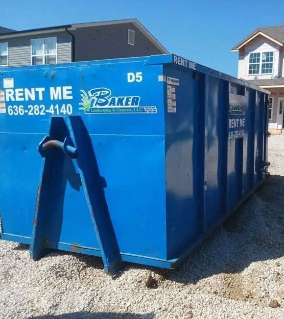 Blue construction dumpster with a RENT ME sign on gravel beside houses.