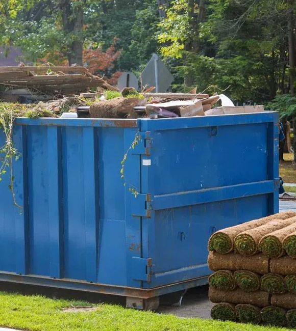 Blue dumpster full of yard debris beside stacked sod rolls in a wooded outdoor area