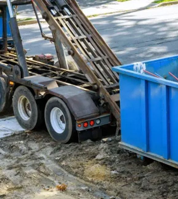 Construction trailer unloading a blue dumpster on a street