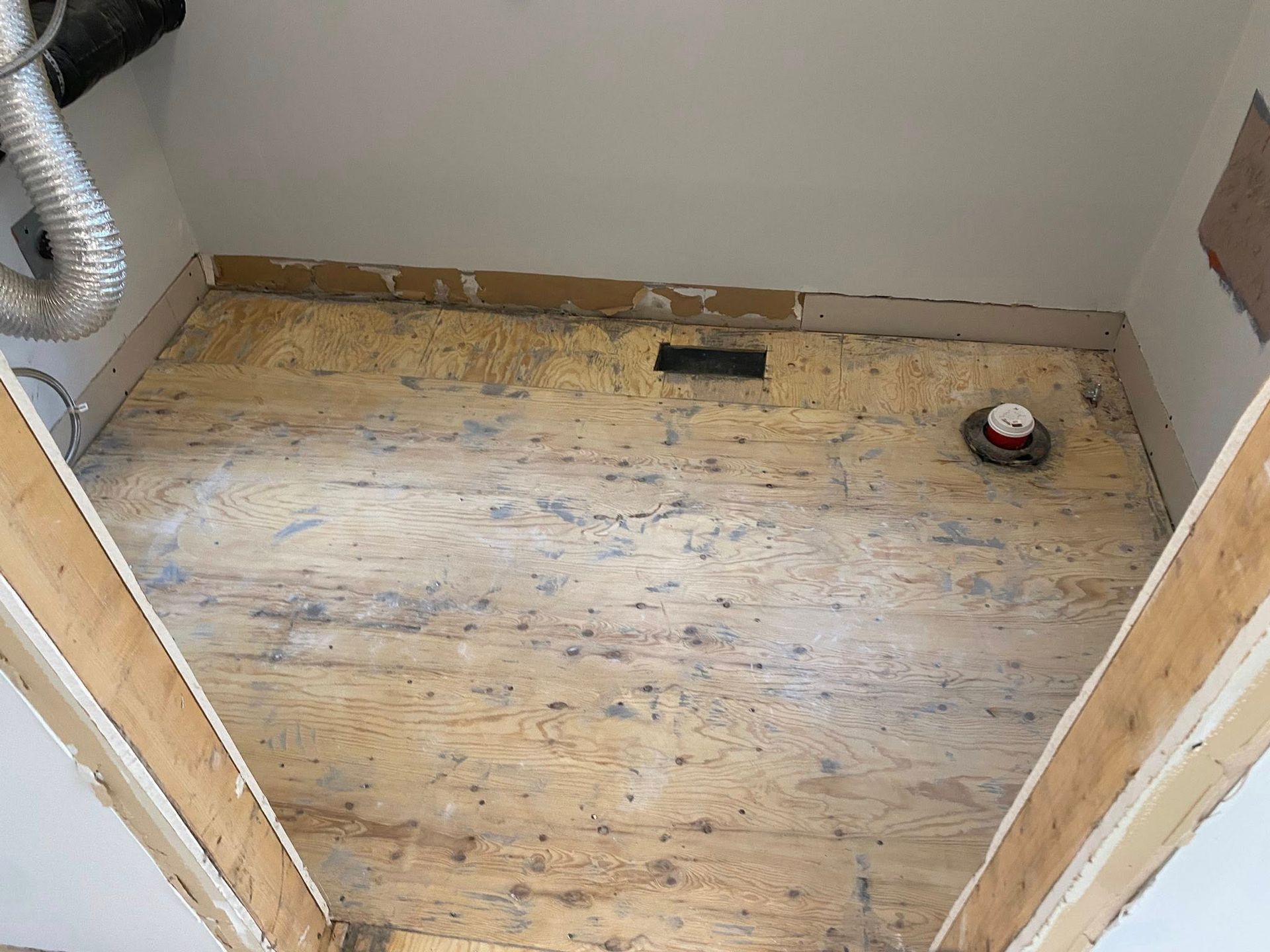 Petite pièce inachevée avec un sous-plancher en bois nu, des conduits apparents et un mètre ruban posé au sol.