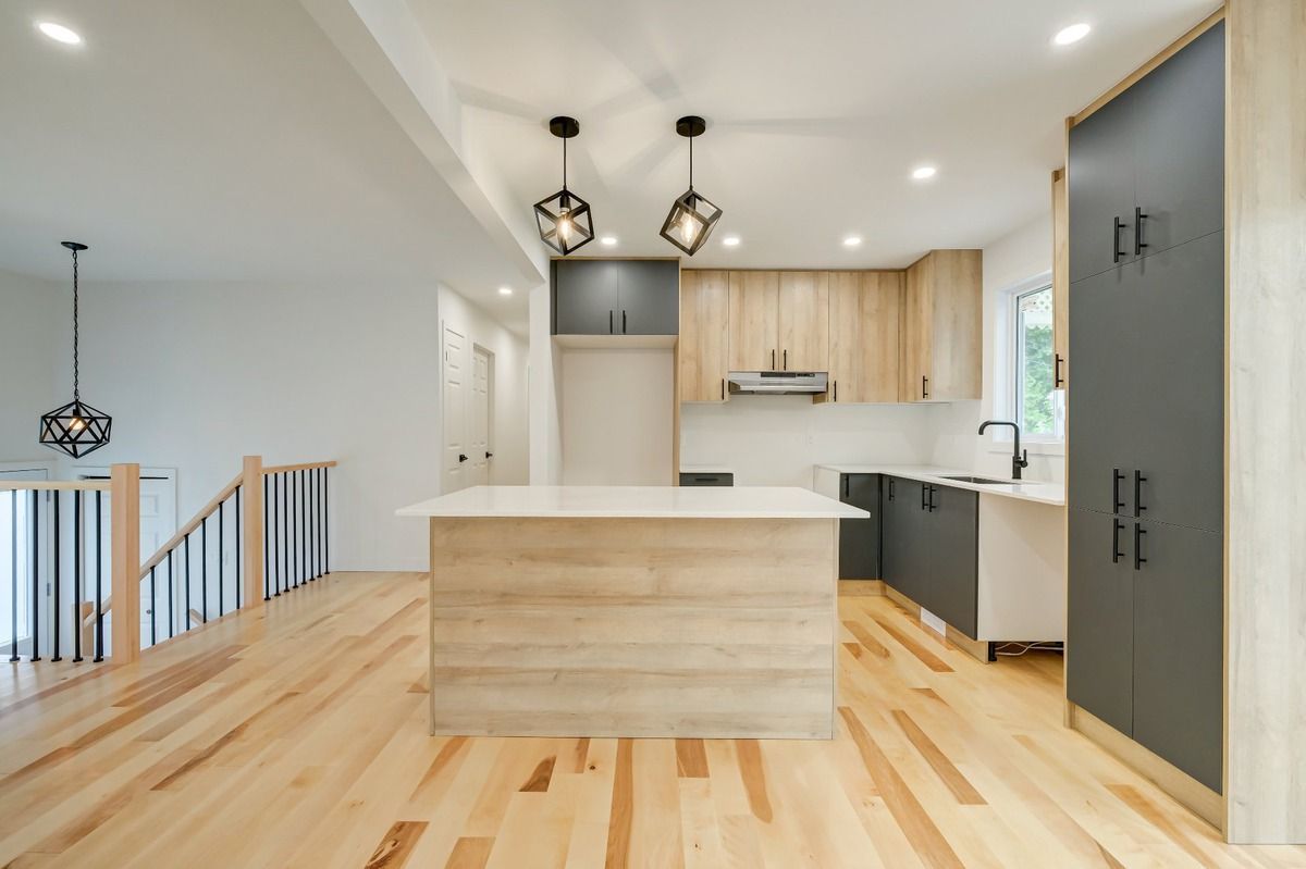 Cuisine moderne et lumineuse avec parquet clair, îlot central blanc, placards gris et suspensions lumineuses.