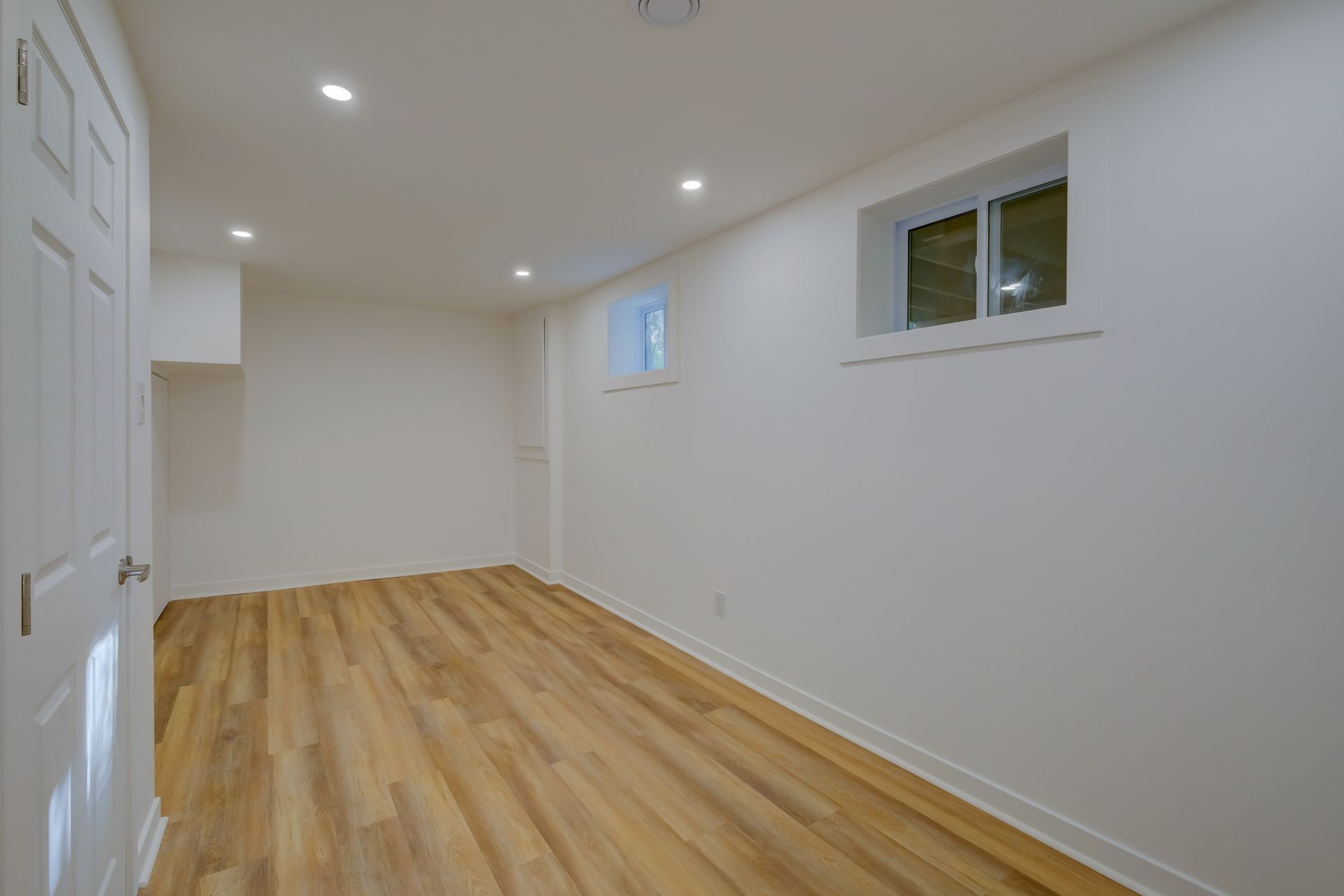 Pièce blanche vide avec parquet, spots encastrés et deux petites fenêtres.