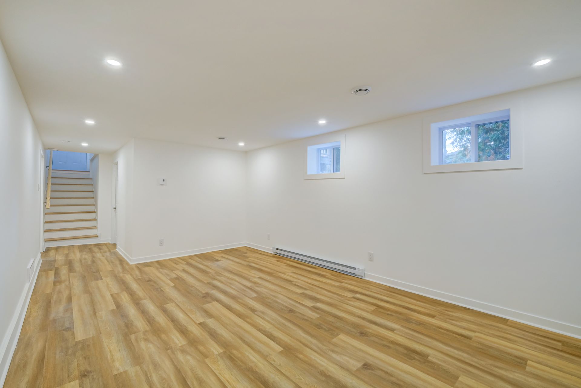 Sous-sol aménagé vide avec parquet, murs blancs, éclairage encastré et petites fenêtres.
