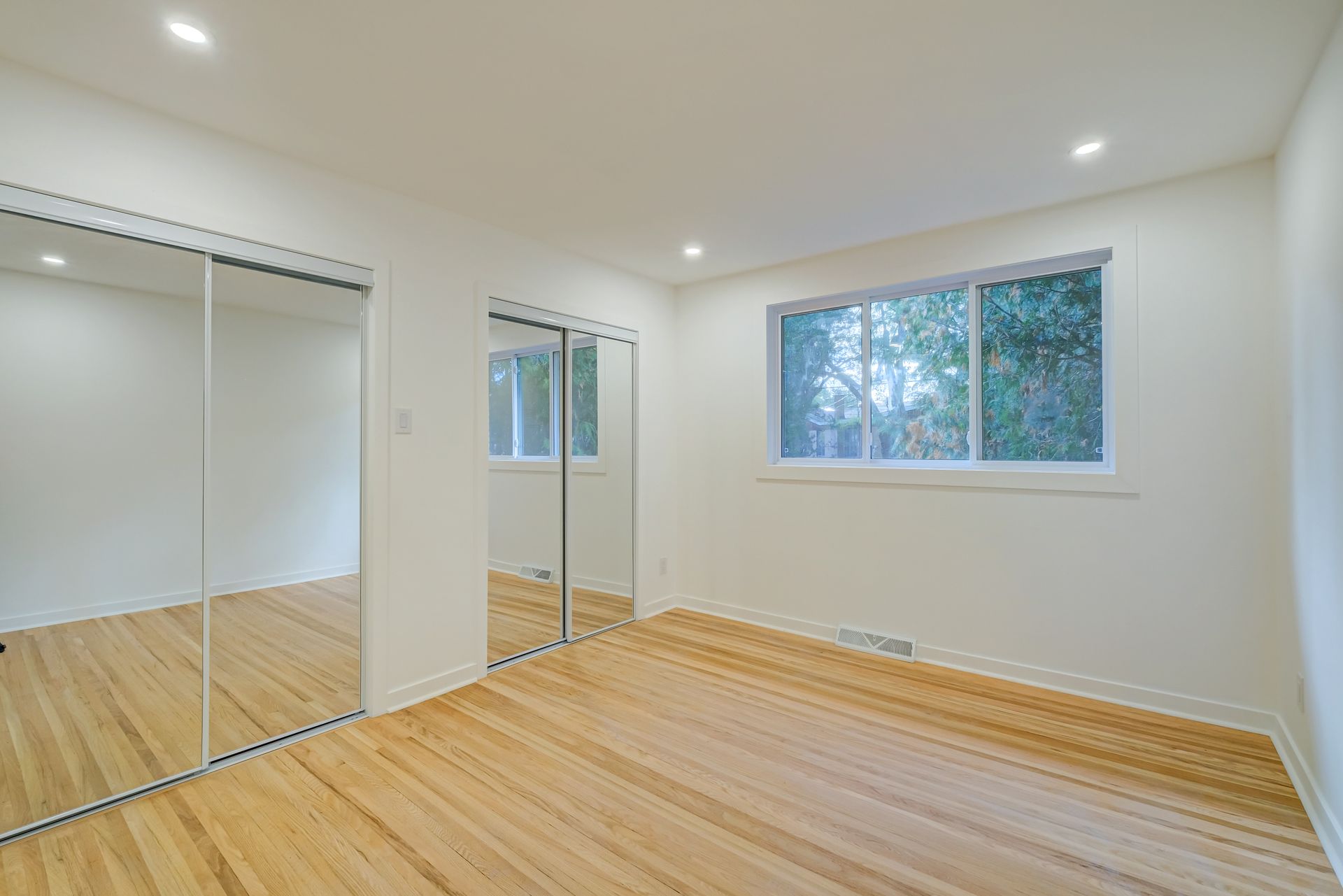 Chambre lumineuse et vide avec parquet, portes de placard à miroir et fenêtre donnant sur les arbres
