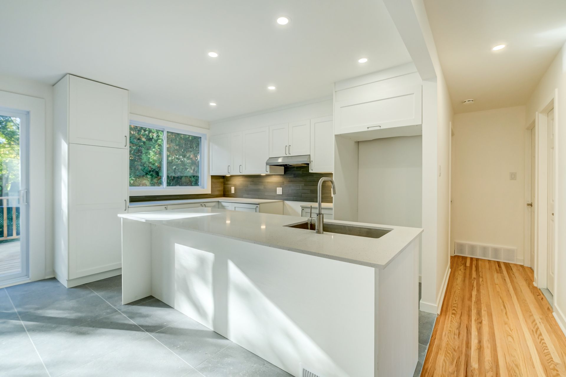Cuisine moderne et lumineuse avec armoires blanches, îlot central, sol gris et couloir en bois attenant.