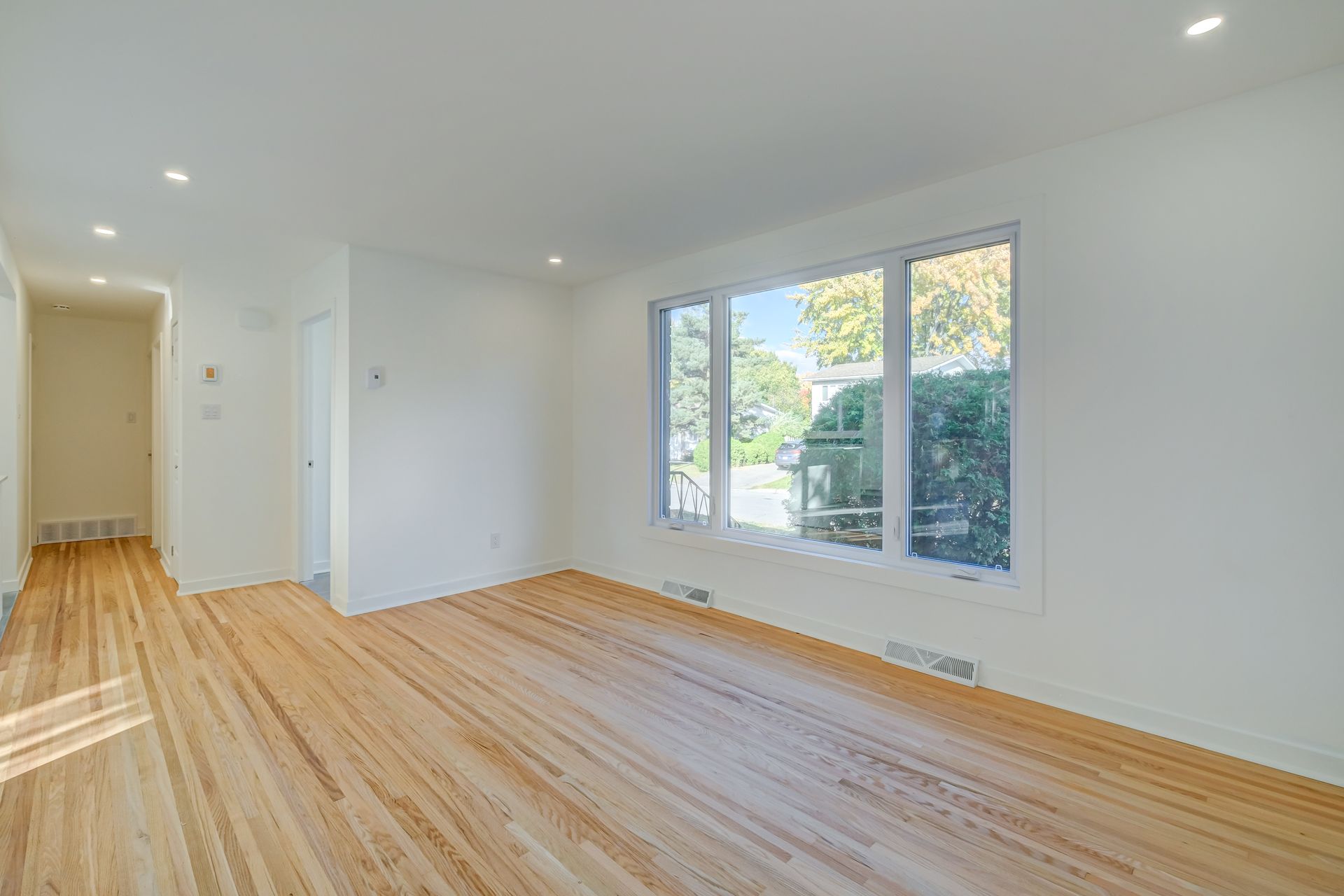 Pièce lumineuse et vide avec parquet, murs blancs, éclairage encastré et grandes fenêtres donnant sur la verdure.