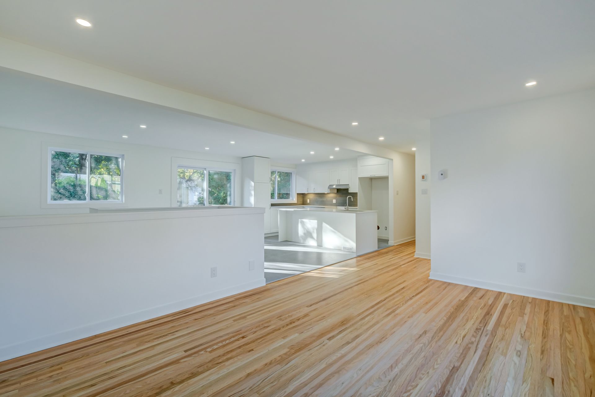 Pièce moderne lumineuse et vide avec parquet, murs blancs, ouverture sur la cuisine et plusieurs petites fenêtres