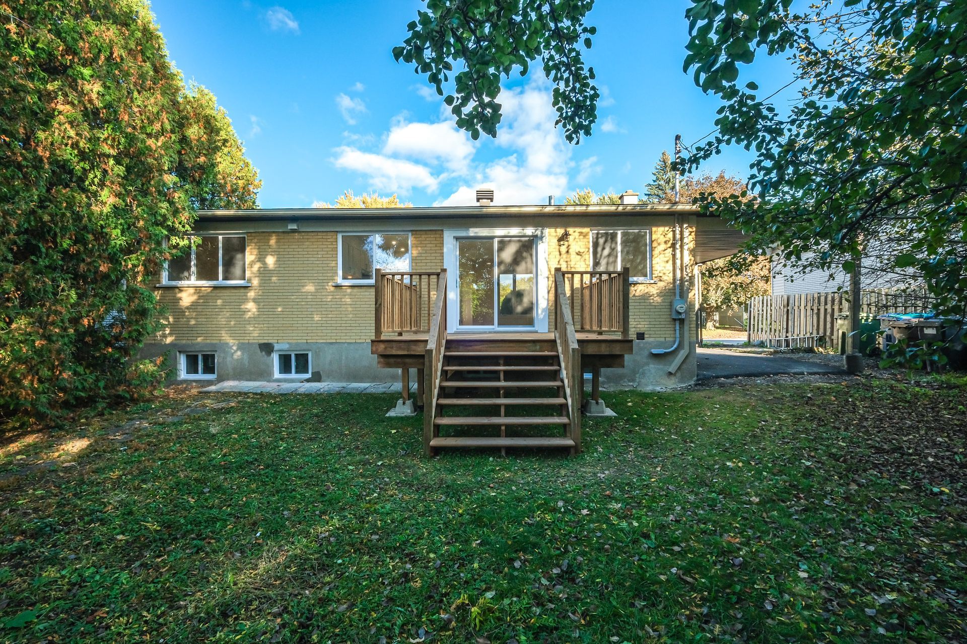 Maison de plain-pied avec terrasse et escalier en bois, entourée d'un jardin arboré sous un ciel bleu.