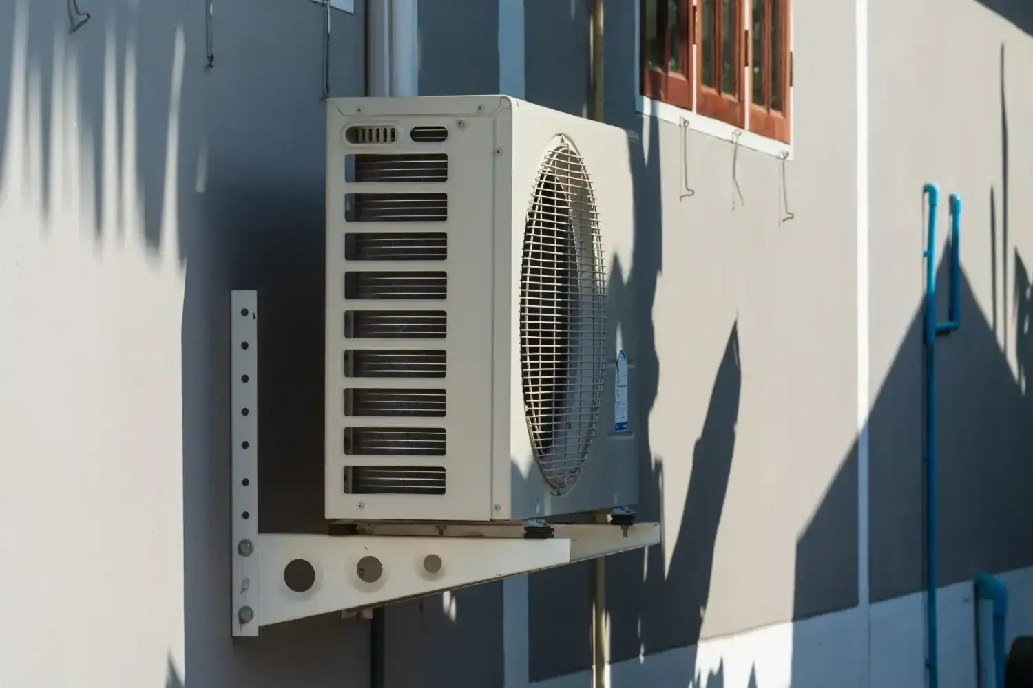 Outdoor air conditioning unit mounted on a wall beside a window, with sunlight and shadows.
