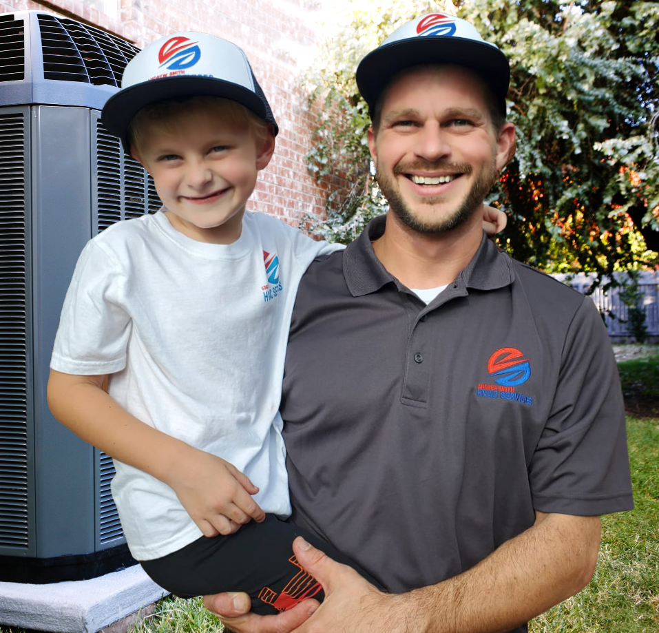 Man holding child, both wearing company hats and shirts, smiling in front of an AC unit.