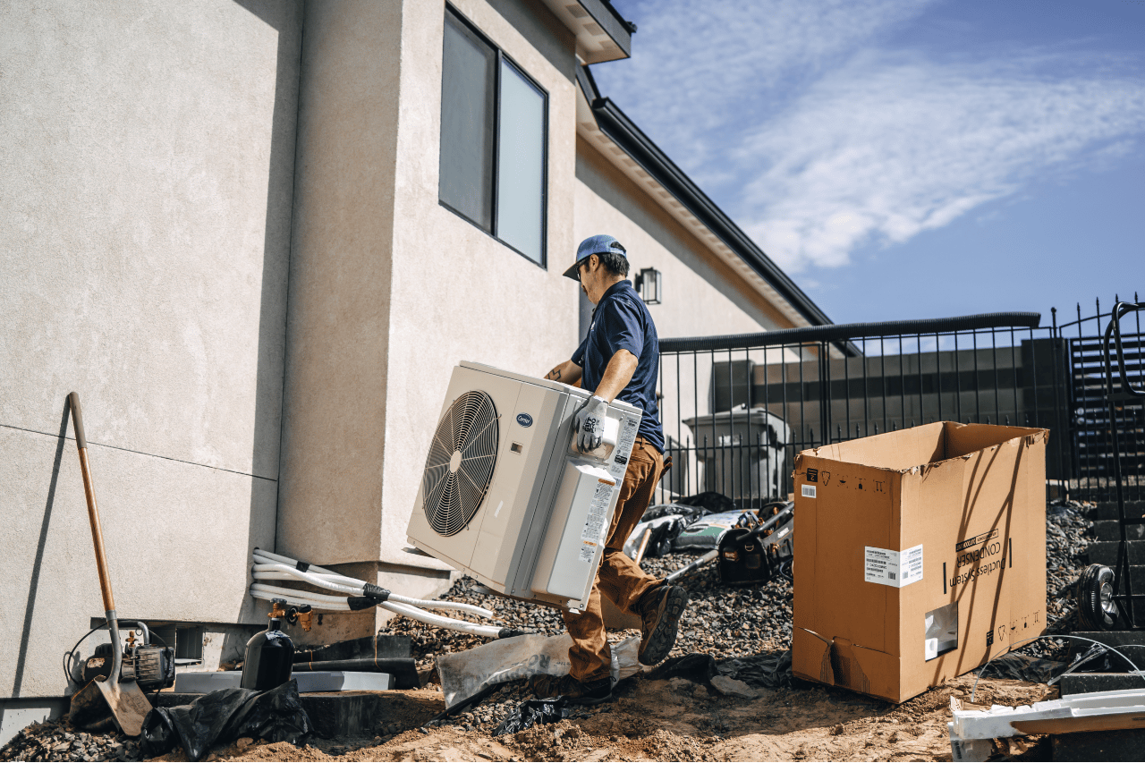 A person carrying an AC unit near a house, with a box and debris visible.