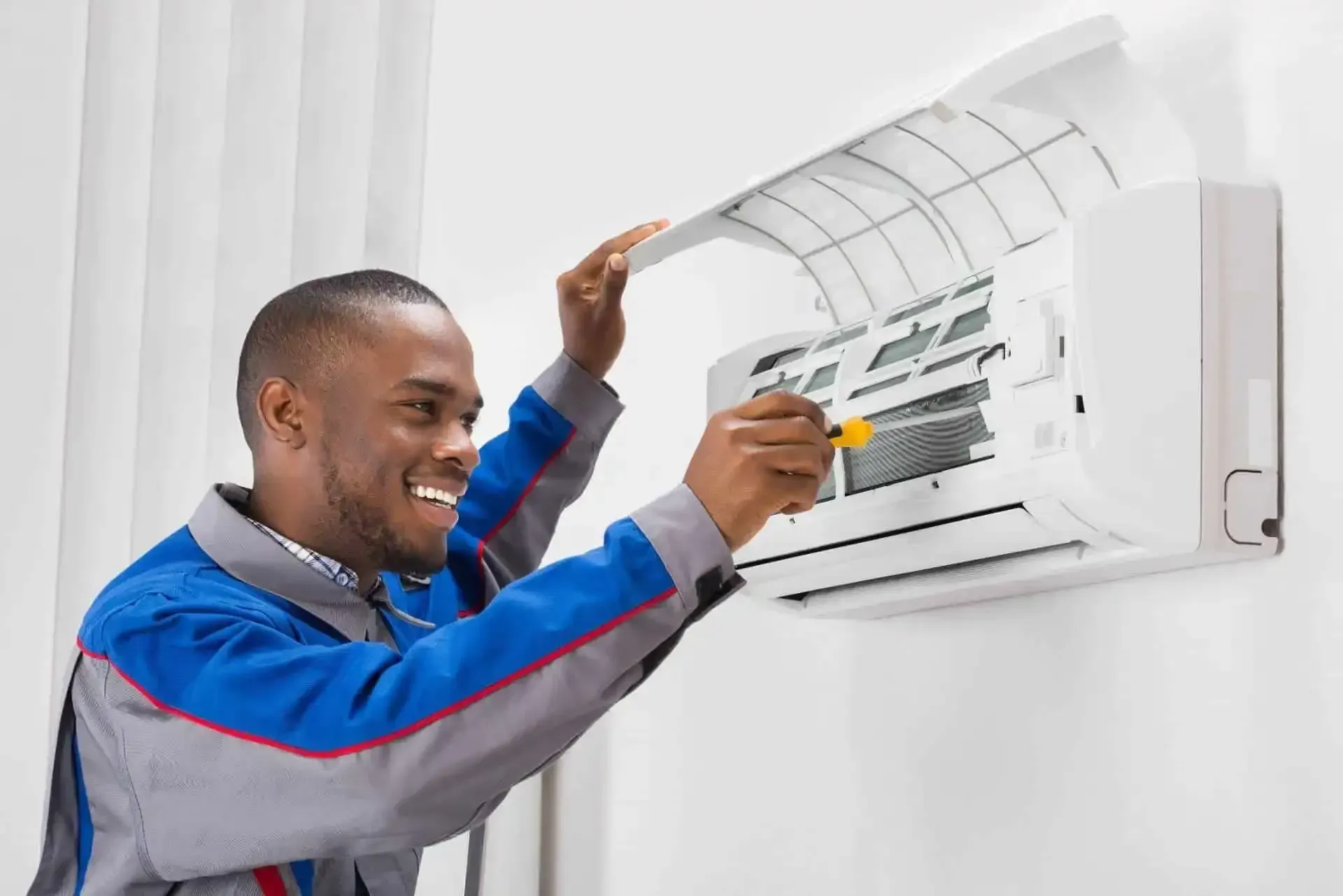 Man in blue jumpsuit cleaning an air conditioner filter with a screwdriver, smiling.