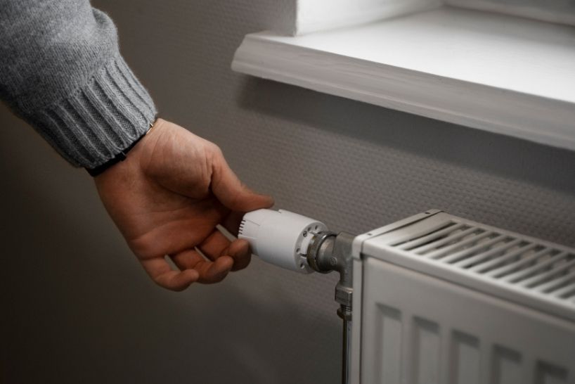 Hand turning the dial of a white radiator next to a window.