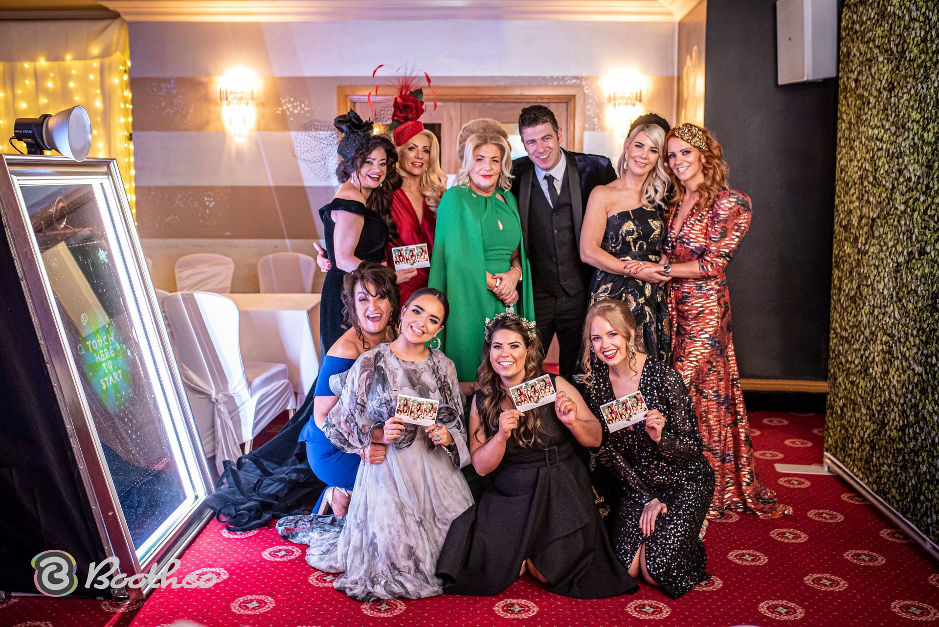 A group of people posing for a photo at a festive event in front of a mirror photo booth on a red patterned carpet.