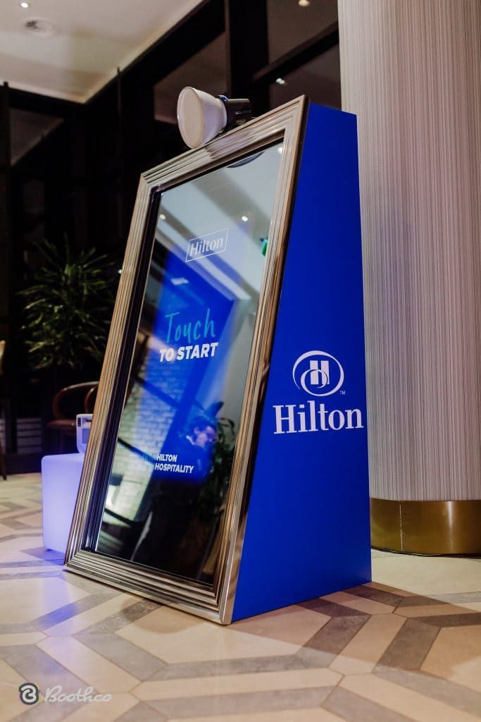 A mirror photo booth with a Hilton logo on its blue side panel, set up in a brightly lit, modern hotel lobby.