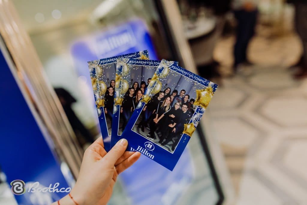 A hand holds three printed photo booth strips featuring a group of people, with blue, gold, and star-themed borders.