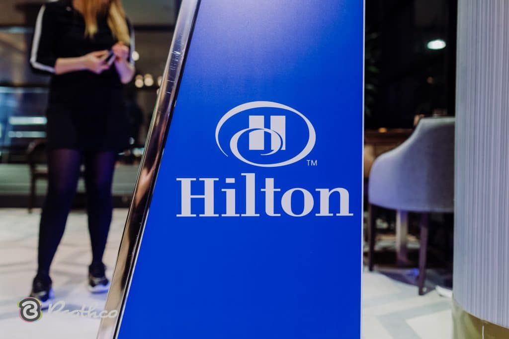 A close-up of a blue sign with the white Hilton logo, with a person in a black outfit visible in the blurred background.