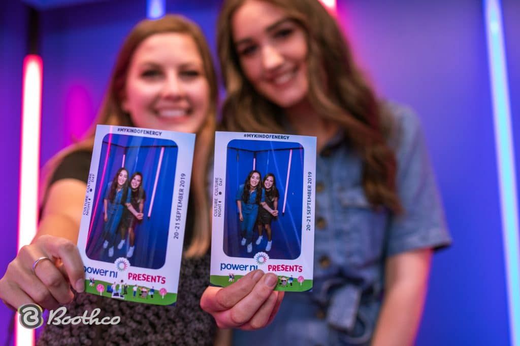 Two people smiling and holding up two matching photo booth prints against a background of purple and blue neon lights.