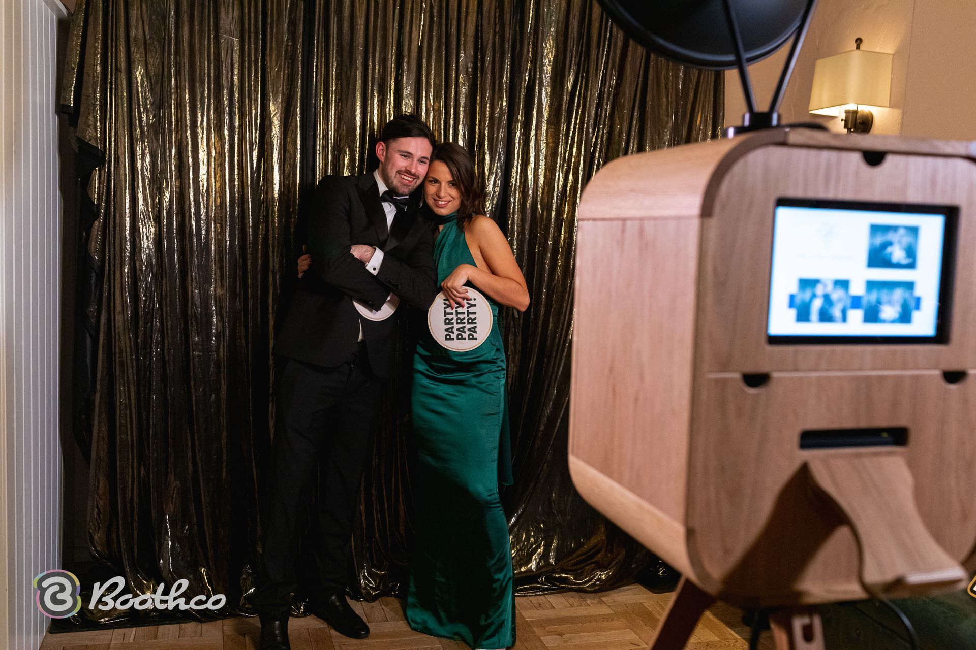 A couple poses together in formal wear in front of a gold sequin curtain for a wooden photo booth.