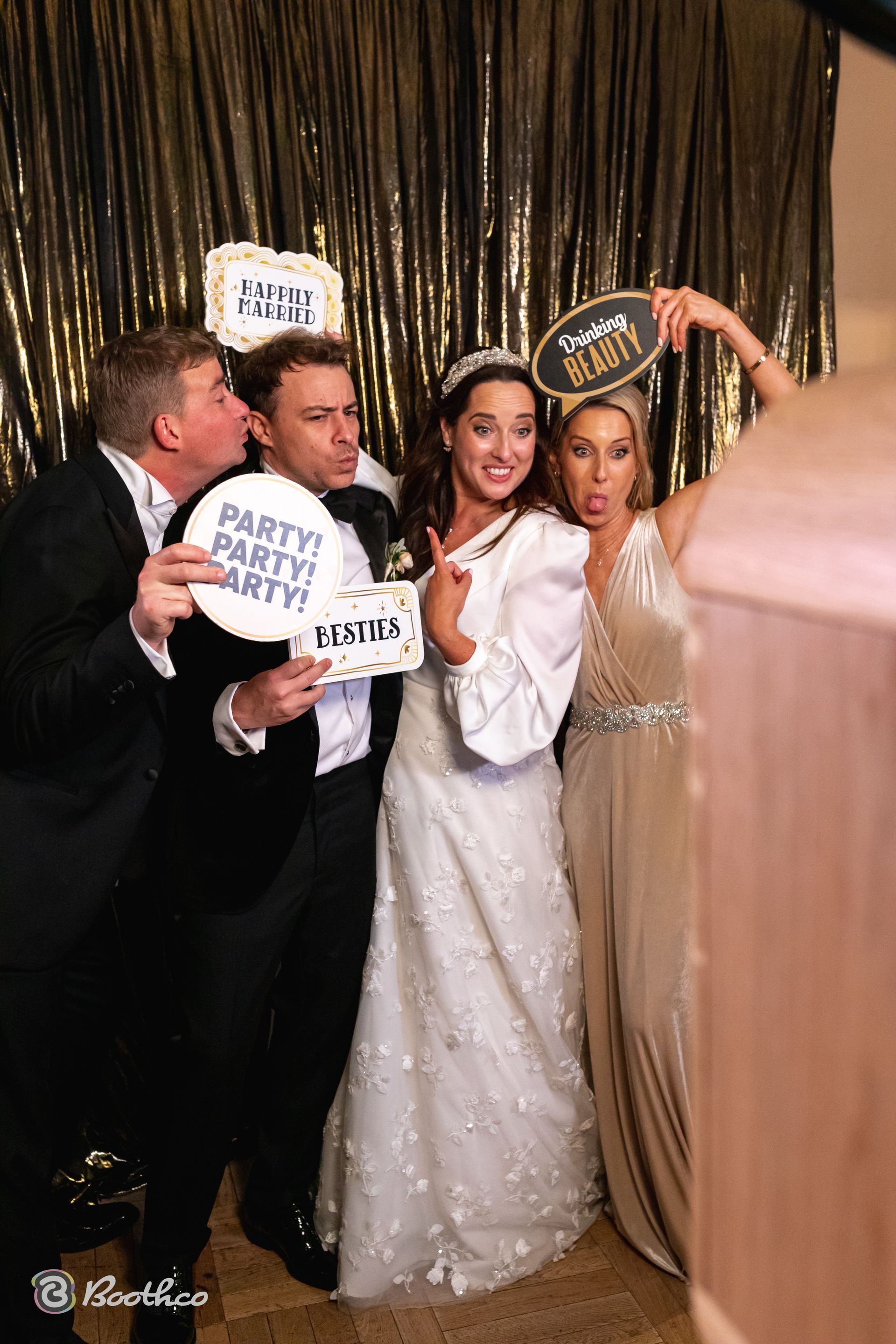 Four people posing in a photo booth with signs, standing in front of a shimmery gold curtain.