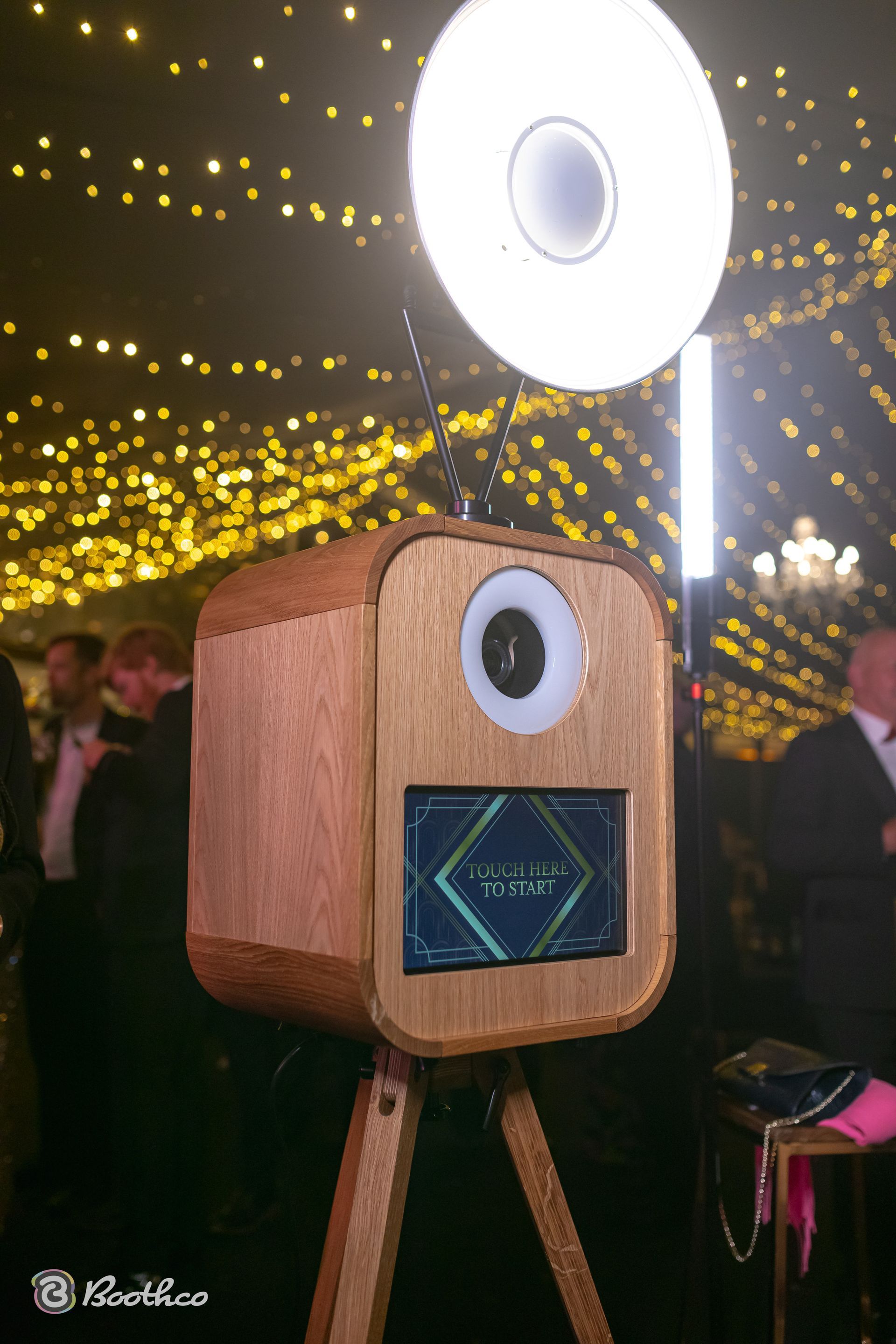 A wooden photo booth with a circular ring light atop a tripod, set up at an evening event with glowing overhead lights.