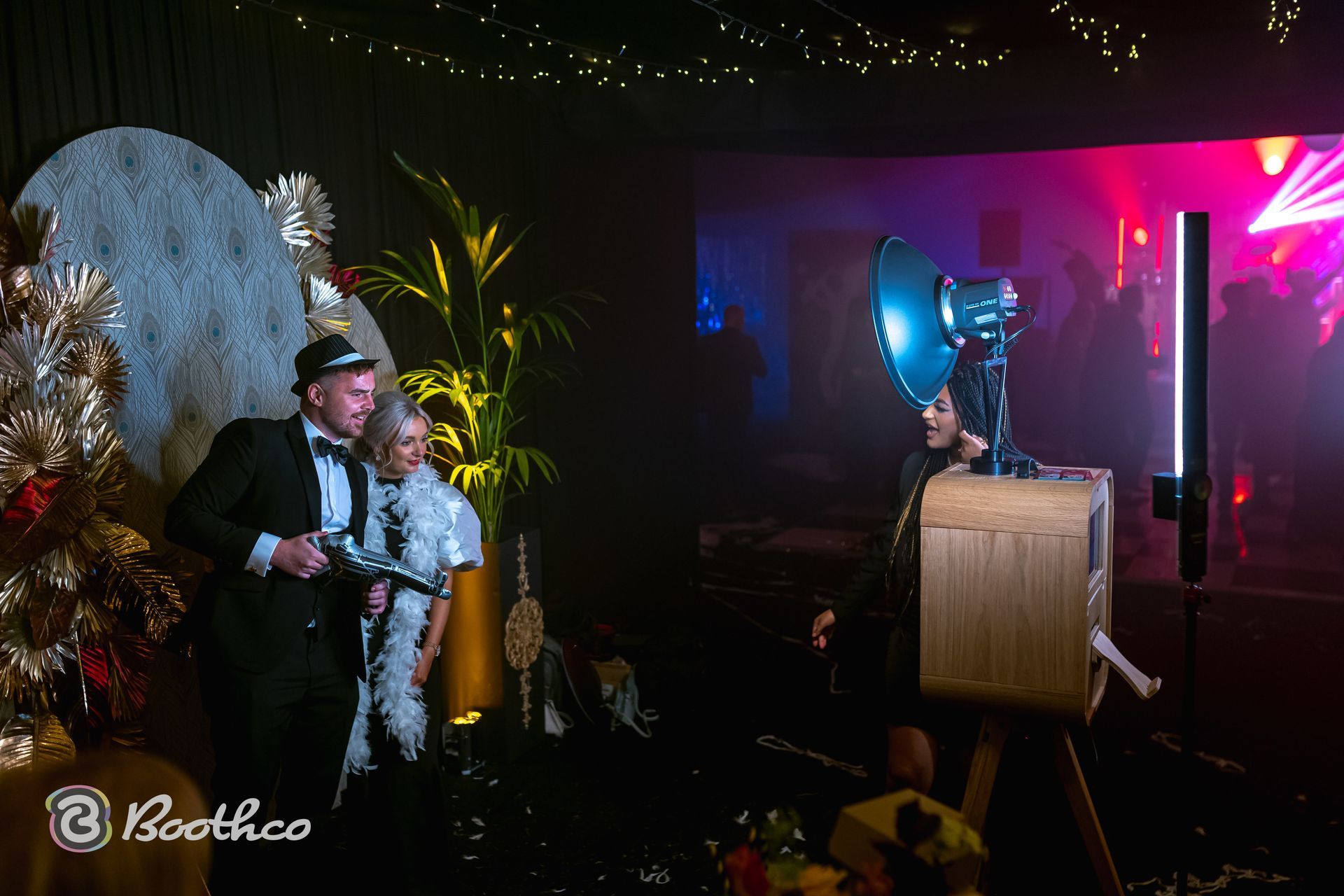 A couple dressed in formal wear poses for a wooden photo booth at a dimly lit, festive evening event.