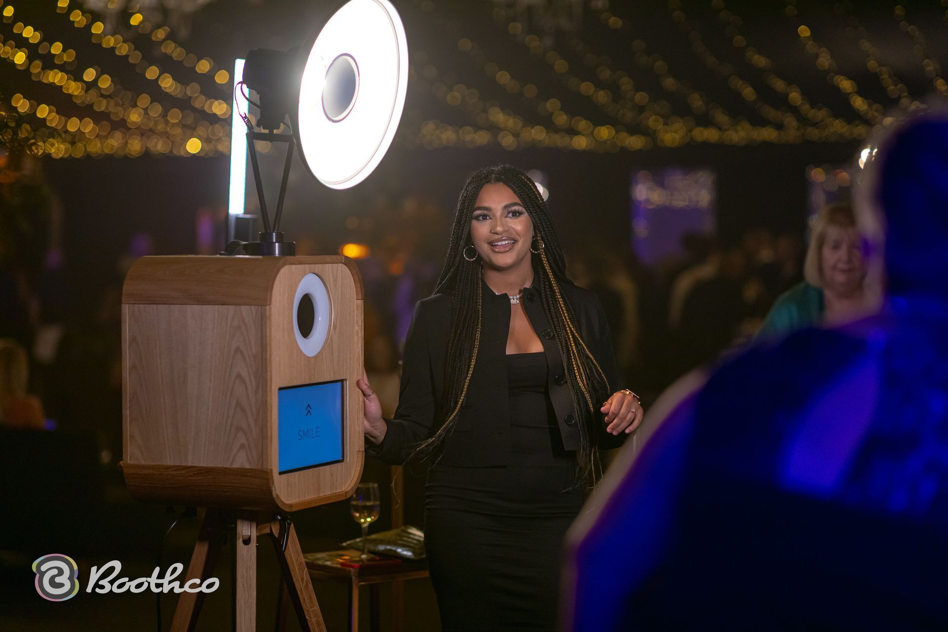 A person smiles while standing next to a wooden photo booth with a ring light at an evening event with fairy lights.