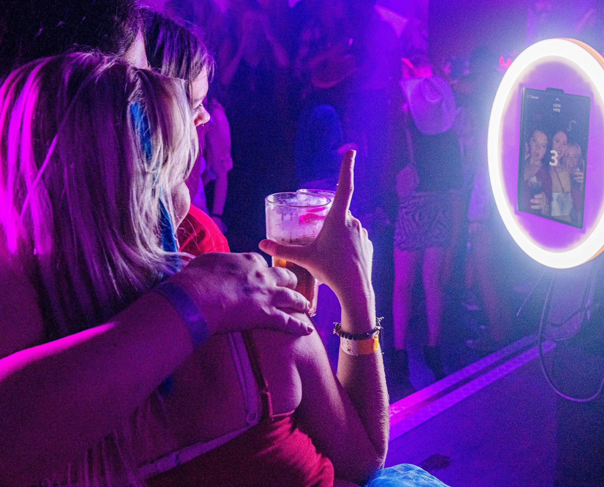 Two people in a dimly lit, purple-hued venue pose for a photo booth while holding drinks.