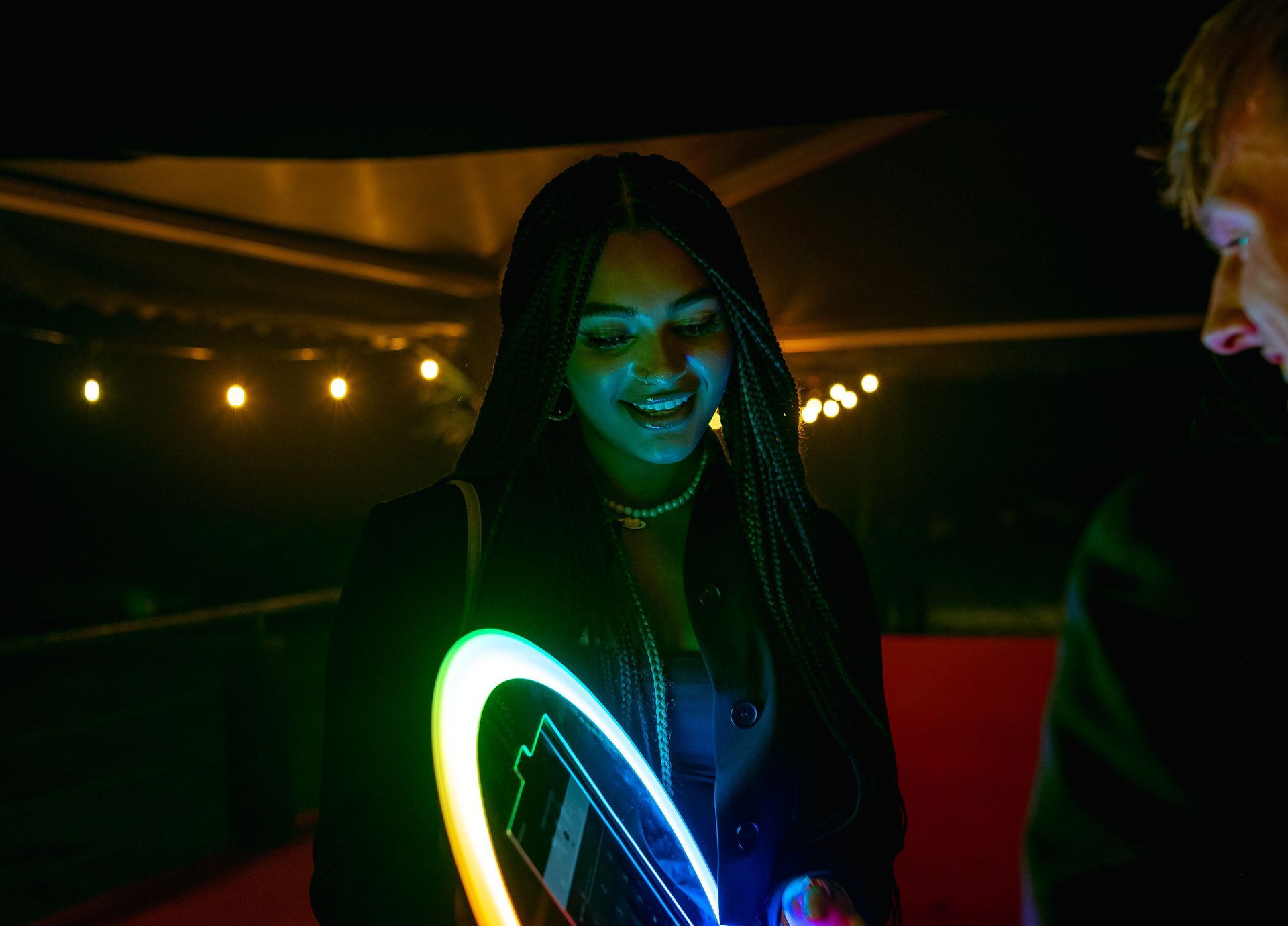 A person smiling while holding a glowing ring light at night, with string lights softly blurred in the background.