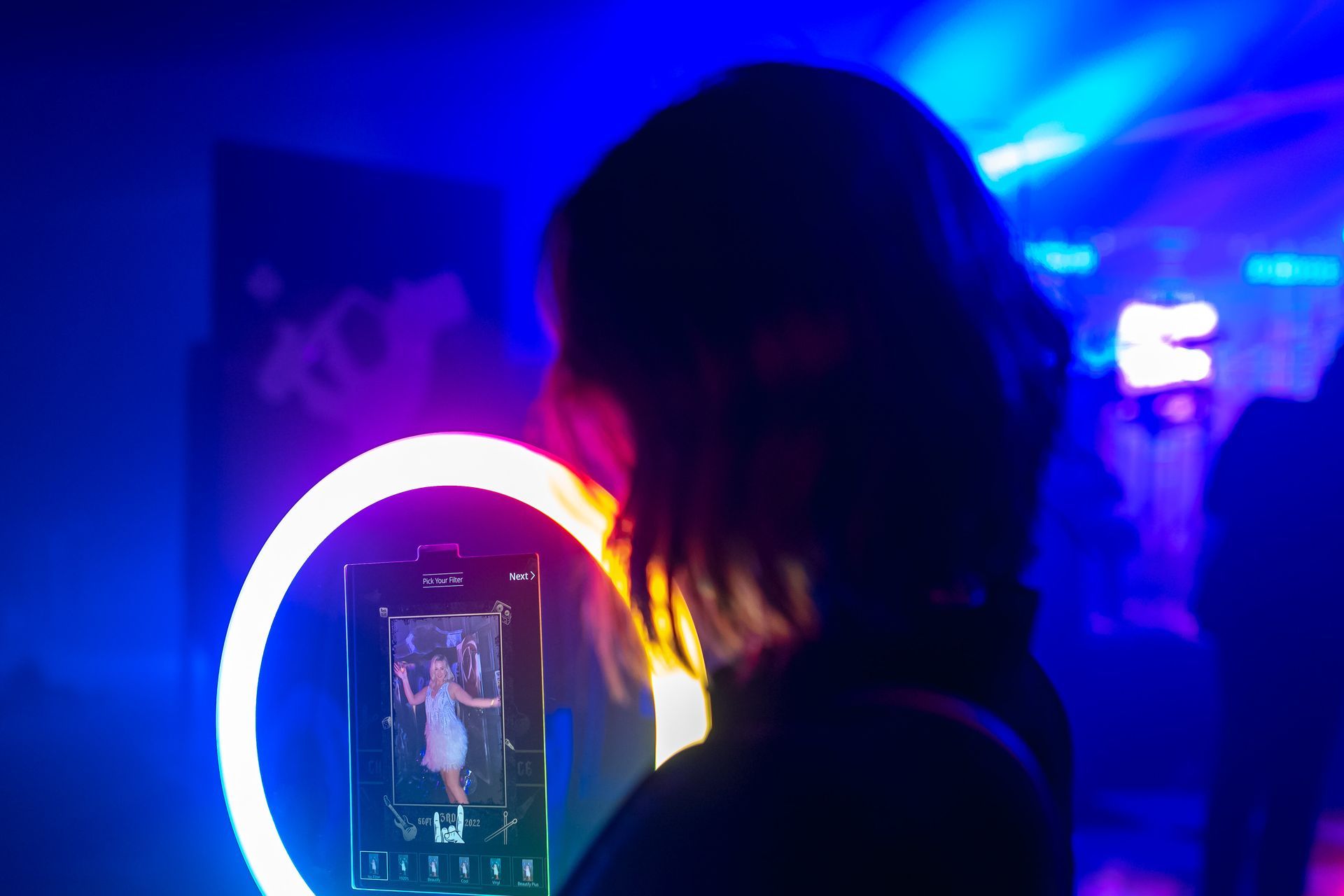 A person in a dim, blue-lit room stands before a glowing ring light and a phone screen displaying a person in a dress.