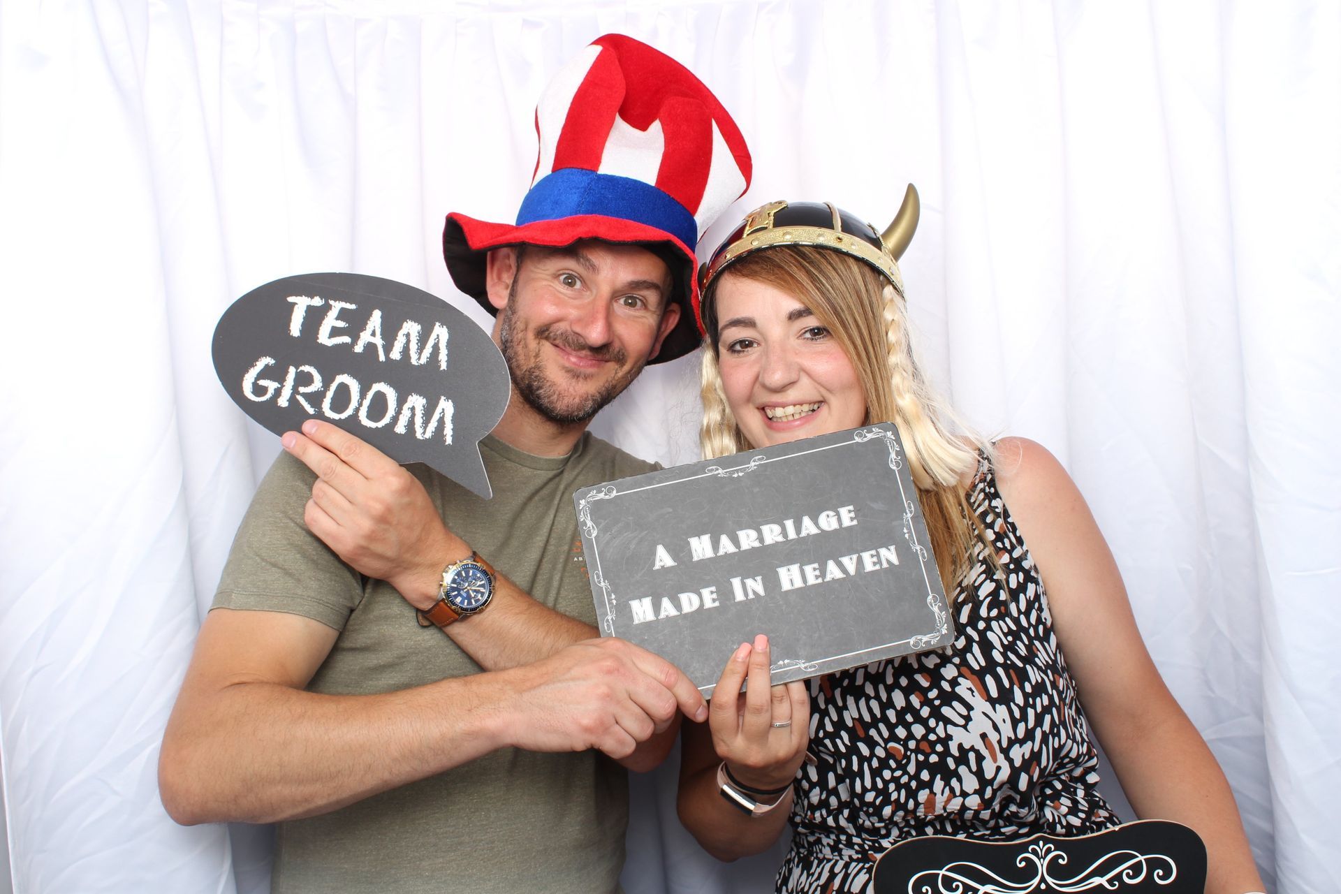 A man and woman pose in a photo booth, wearing fun hats and holding props that say 
