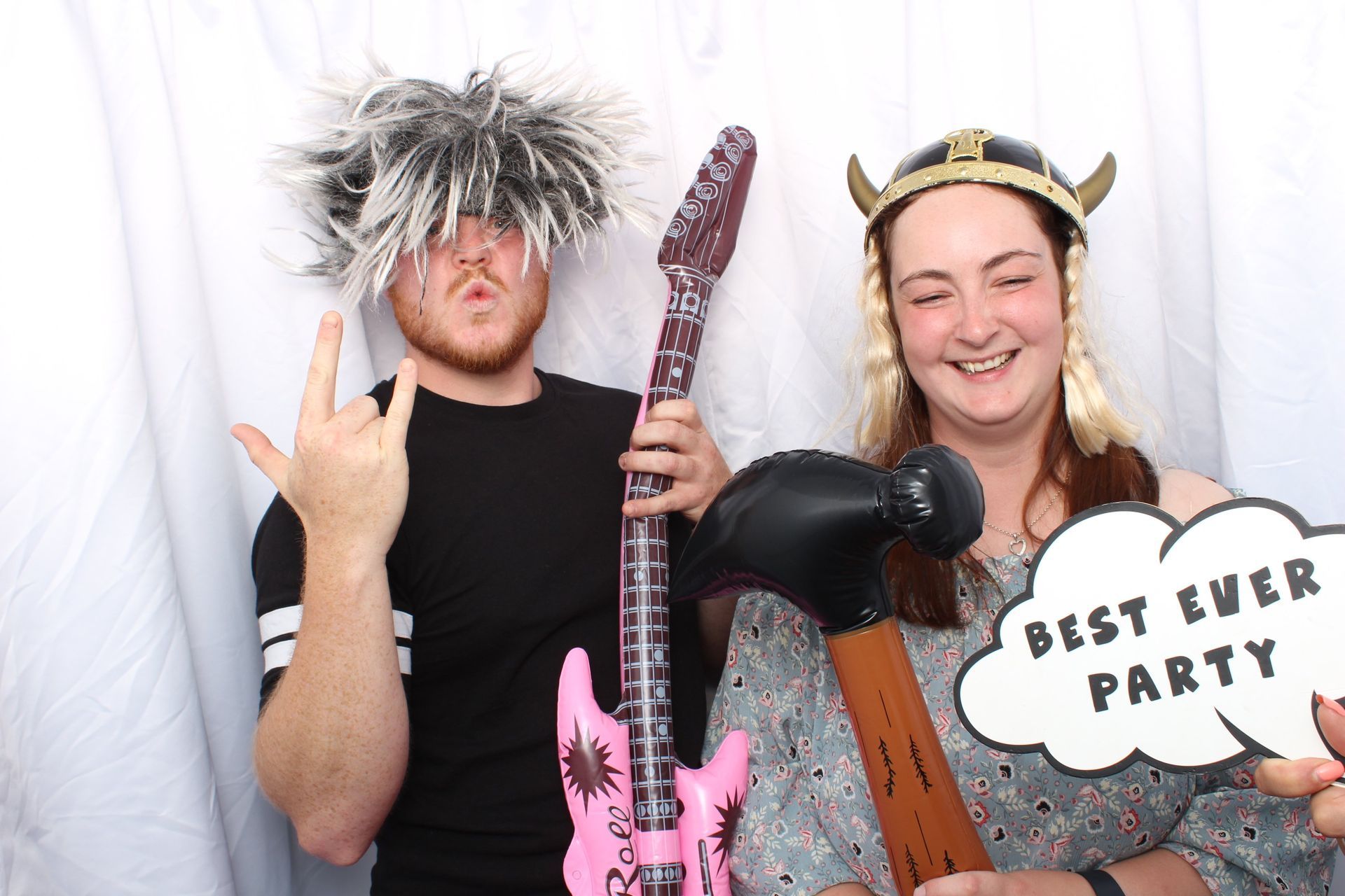 Two people in costumes, one with a spiky wig and inflatable guitar, the other in a Viking helmet holding a party sign.