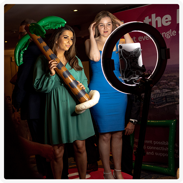 Two people posing for a photo booth with an inflatable palm tree, illuminated by a ring light.