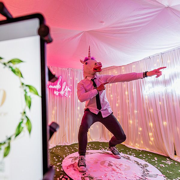 A person wearing a unicorn mask, white shirt, and black tie poses playfully on a lit stage with pink lighting and curtains.