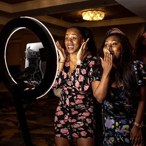 Two people in floral dresses look excitedly at a tablet mounted inside a glowing ring light at an indoor event.