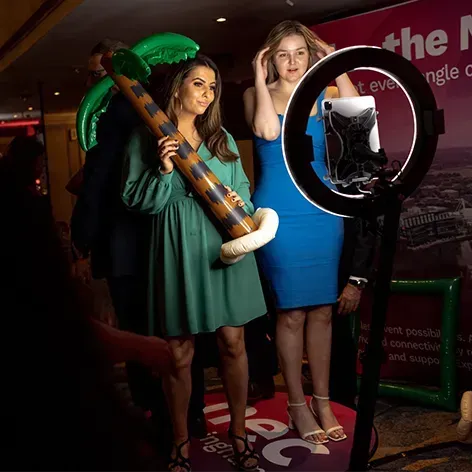 Two people stand before a ring light and tablet on a stand; one holds an inflatable palm tree while the other adjusts hair.