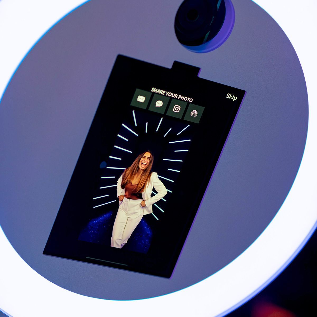 A photo booth screen shows a smiling person with arms on hips, framed by a circular light ring with share icons above.