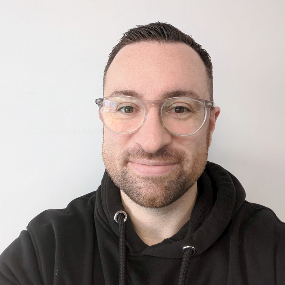 A person with short brown hair, wearing clear-framed glasses and a black hoodie, smiles against a plain white background.