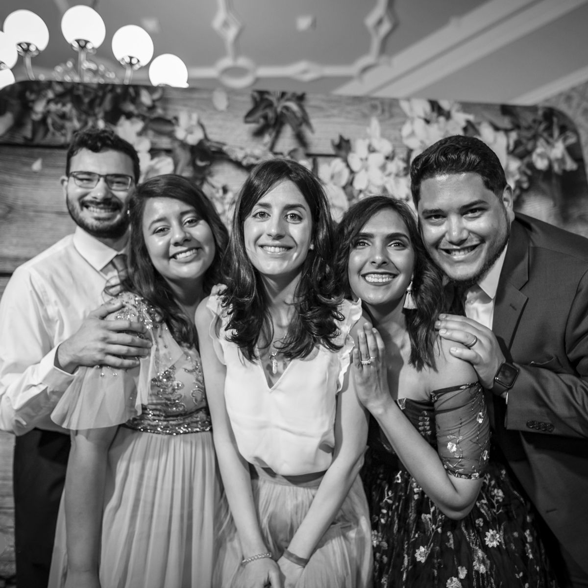 Five people pose together, smiling warmly, in front of a floral backdrop at a formal event, captured in monochrome.