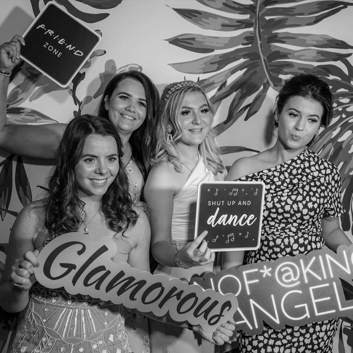 Four people posing with fun photo props against a palm-leaf backdrop in a black-and-white photo booth setting.