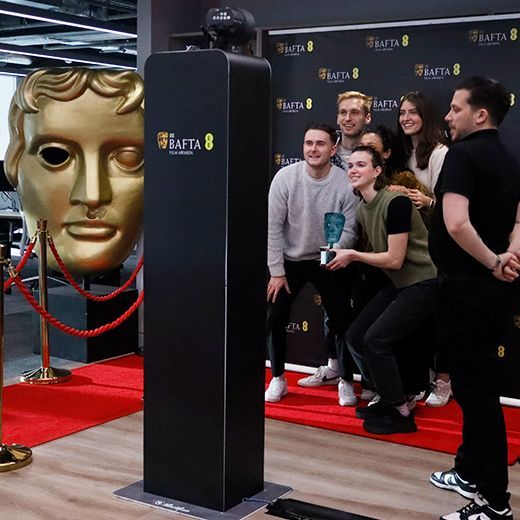 A group celebrates at a BAFTA event, posing for a photo near a large gold BAFTA mask statue and a branded pillar.