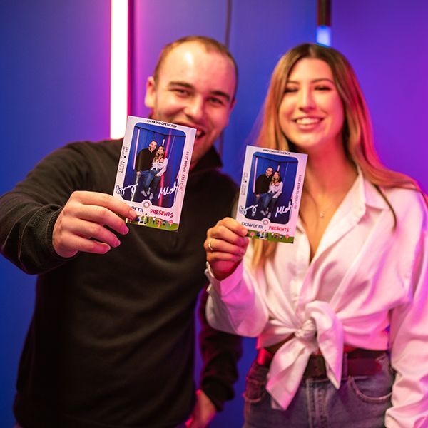 A smiling couple stands in front of a blue background with pink lighting, holding up their photo booth strips.