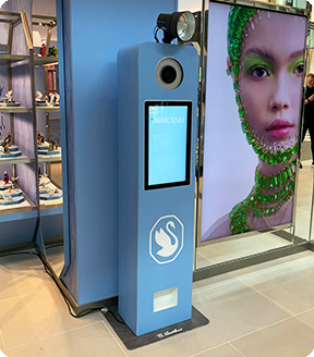A light blue Swarovski-branded photo booth kiosk stands next to a large display featuring a model in green jewelry.