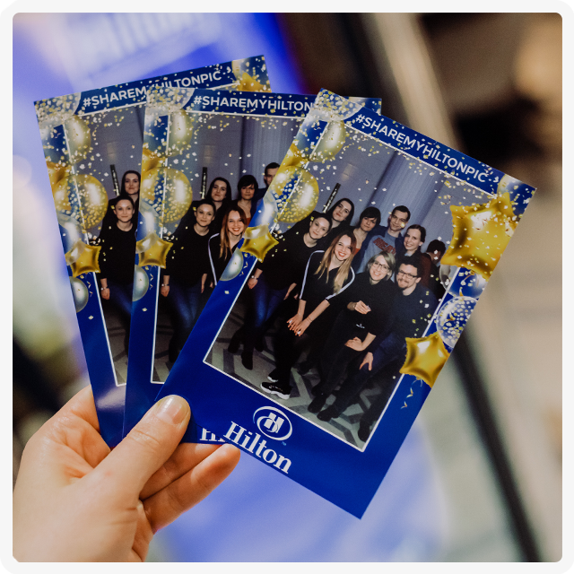 A hand holds three printed photo booth strips from a Hilton event, showing groups of people smiling in front of decor.