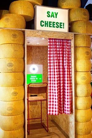 A photo booth framed by stacked wheels of cheese, with a red-and-white checkered curtain and a 
