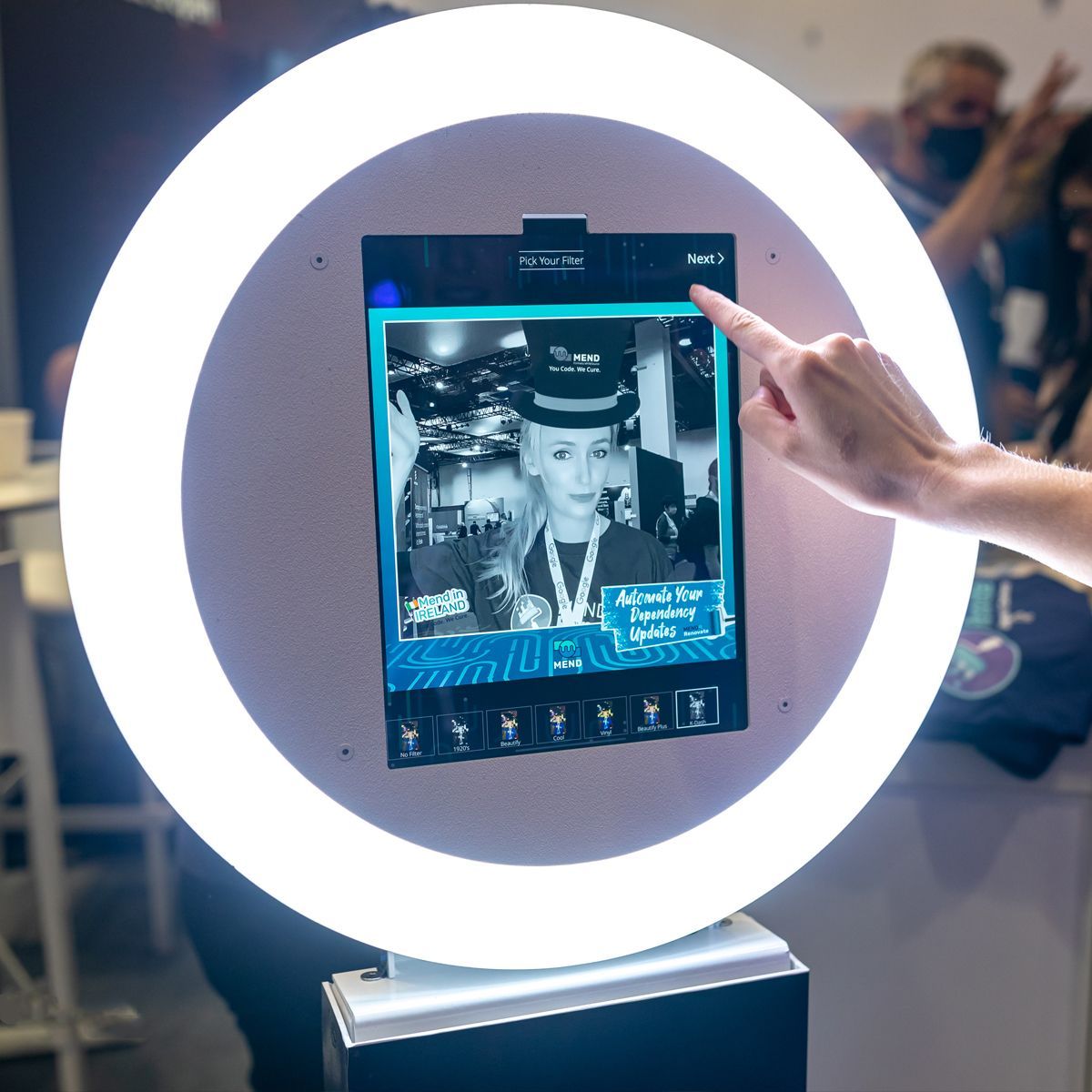 A person's finger touches a digital photo booth screen displaying a black-and-white portrait framed by a bright ring light.
