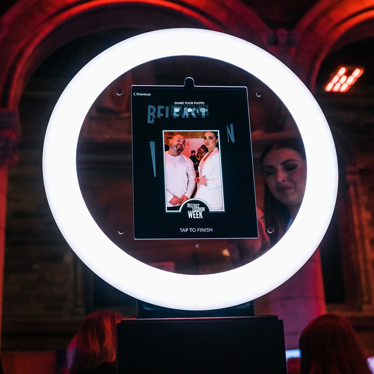 A ring light frames a tablet displaying a photo of two people, with a person visible to the side in a dimly lit setting.