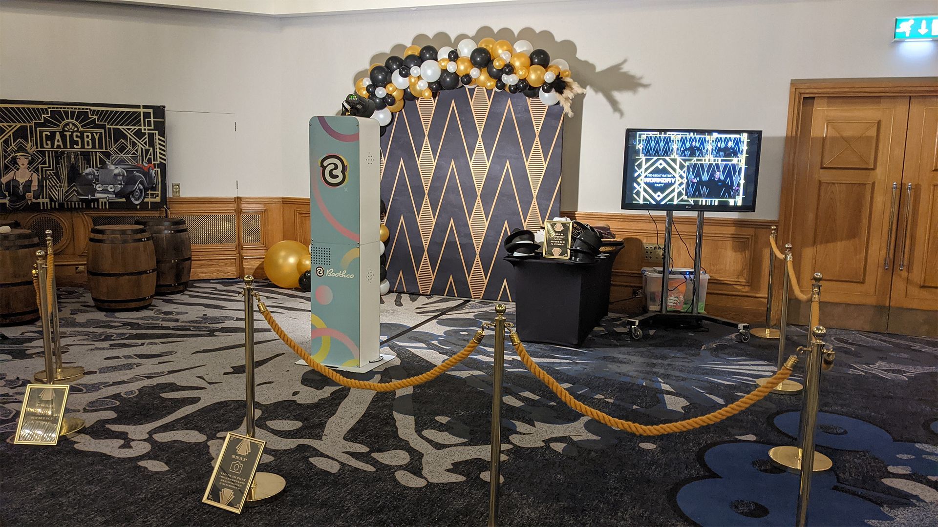 A photo booth setup with a patterned backdrop, balloon arch, signage, screen, and stanchions on a patterned carpet.